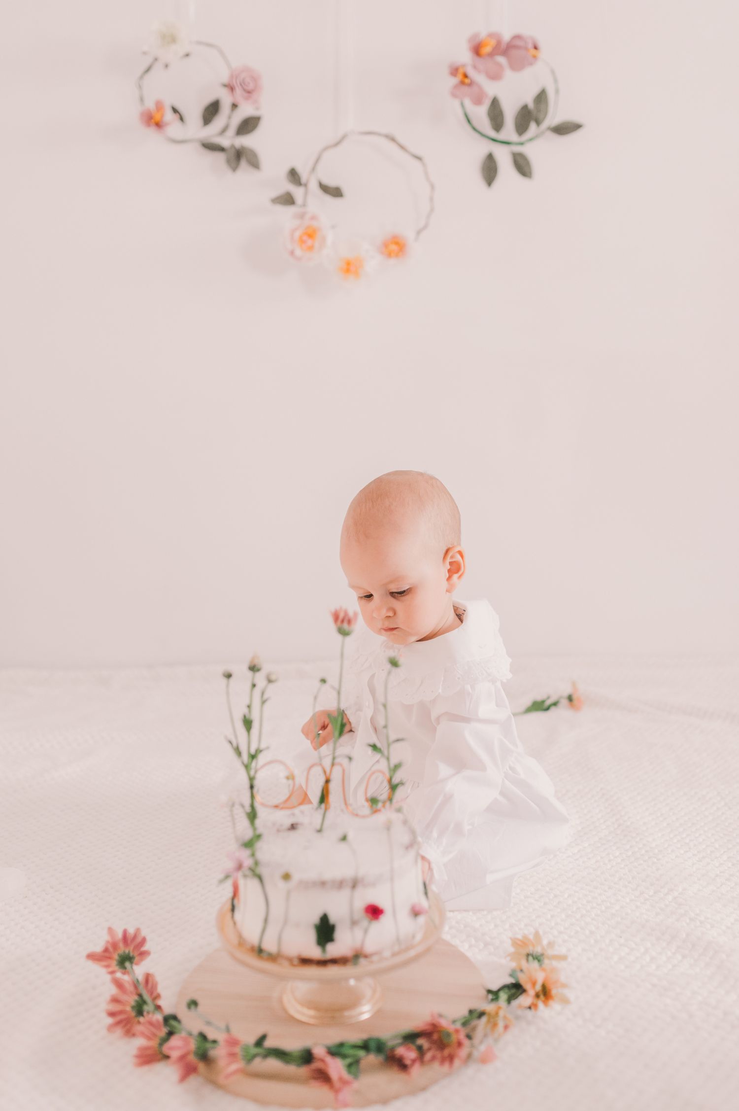 Baby in white dress explores floral decorations in soft pink studio setup.
