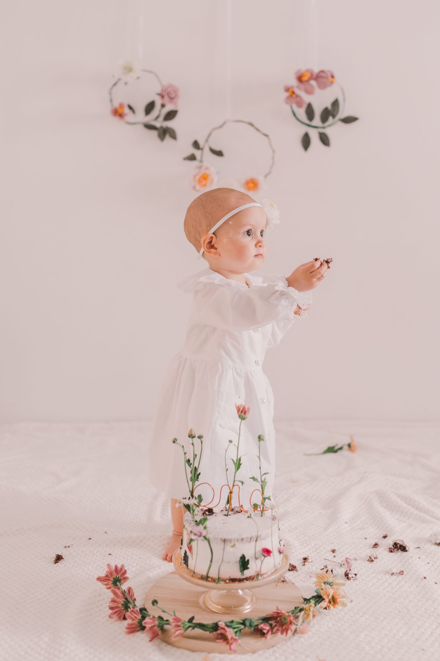 Baby in white dress plays with small flowers in ethereal white studio setting.