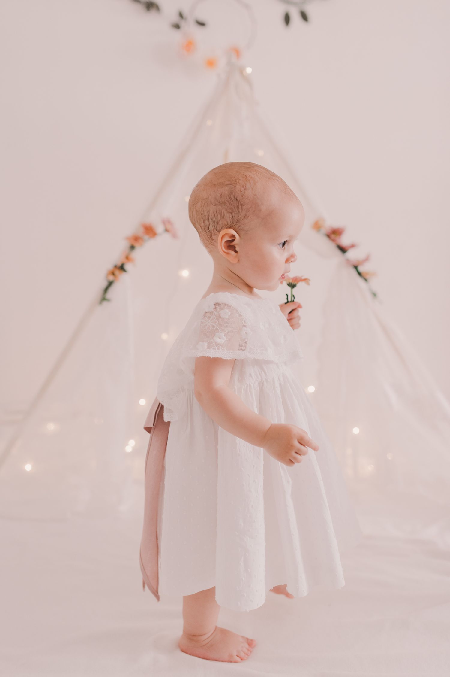 Baby wearing white dress stands near illuminated teepee with fairy lights.