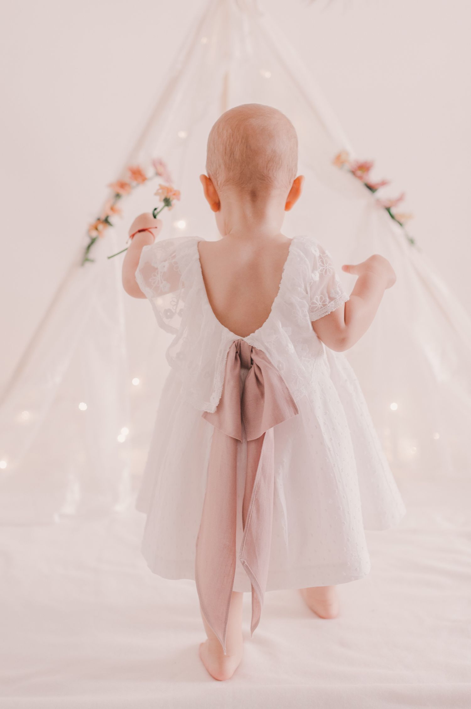 Baby in white dress explores magical teepee setup with twinkling lights.