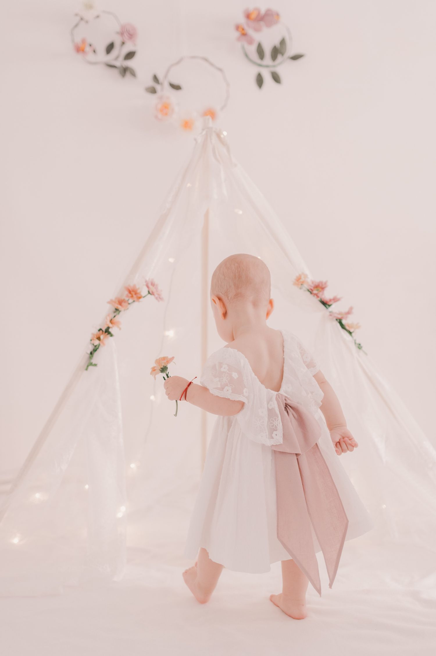Baby in white dress explores whimsical teepee setting with string lights.