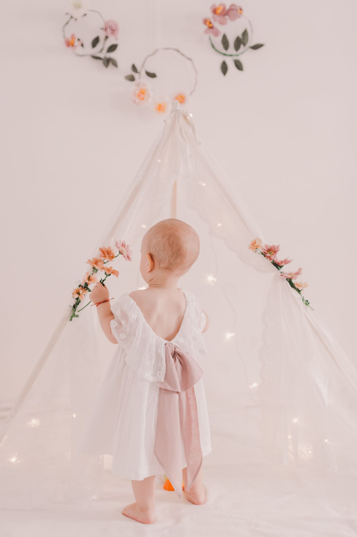 Baby in white dress plays near illuminated teepee with floral decorations.