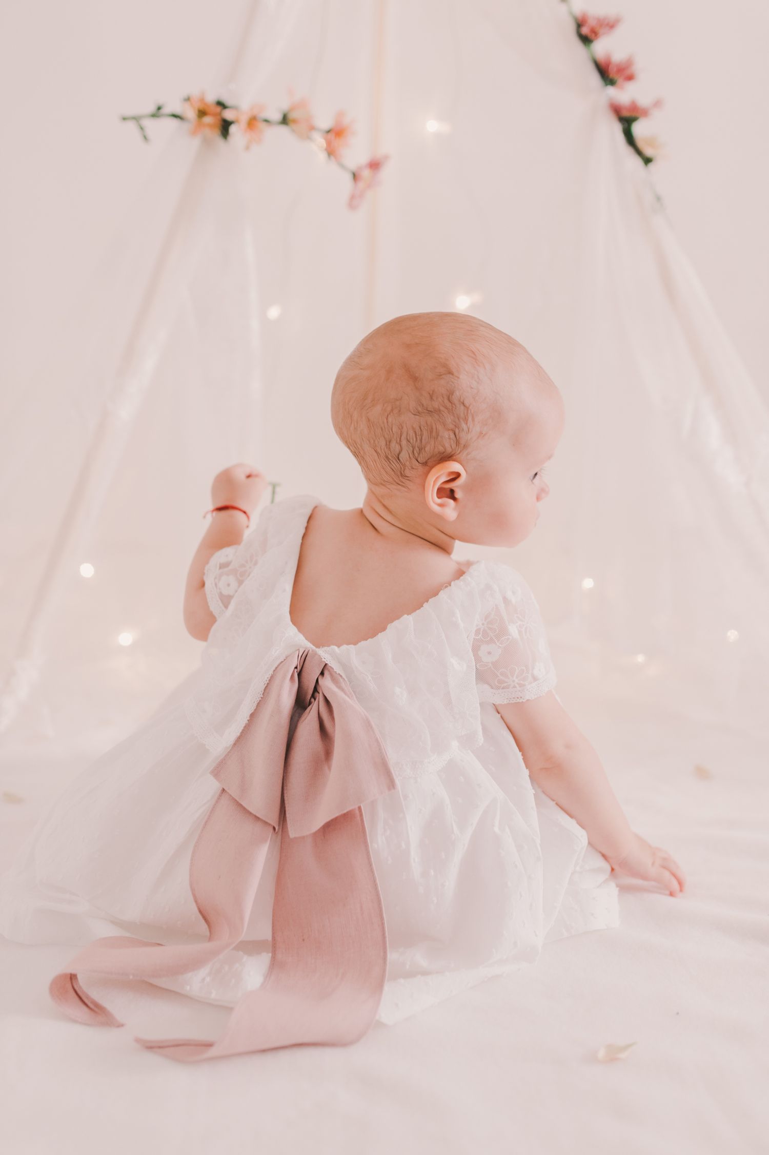 Baby in white dress crawls near decorated teepee with soft lighting.