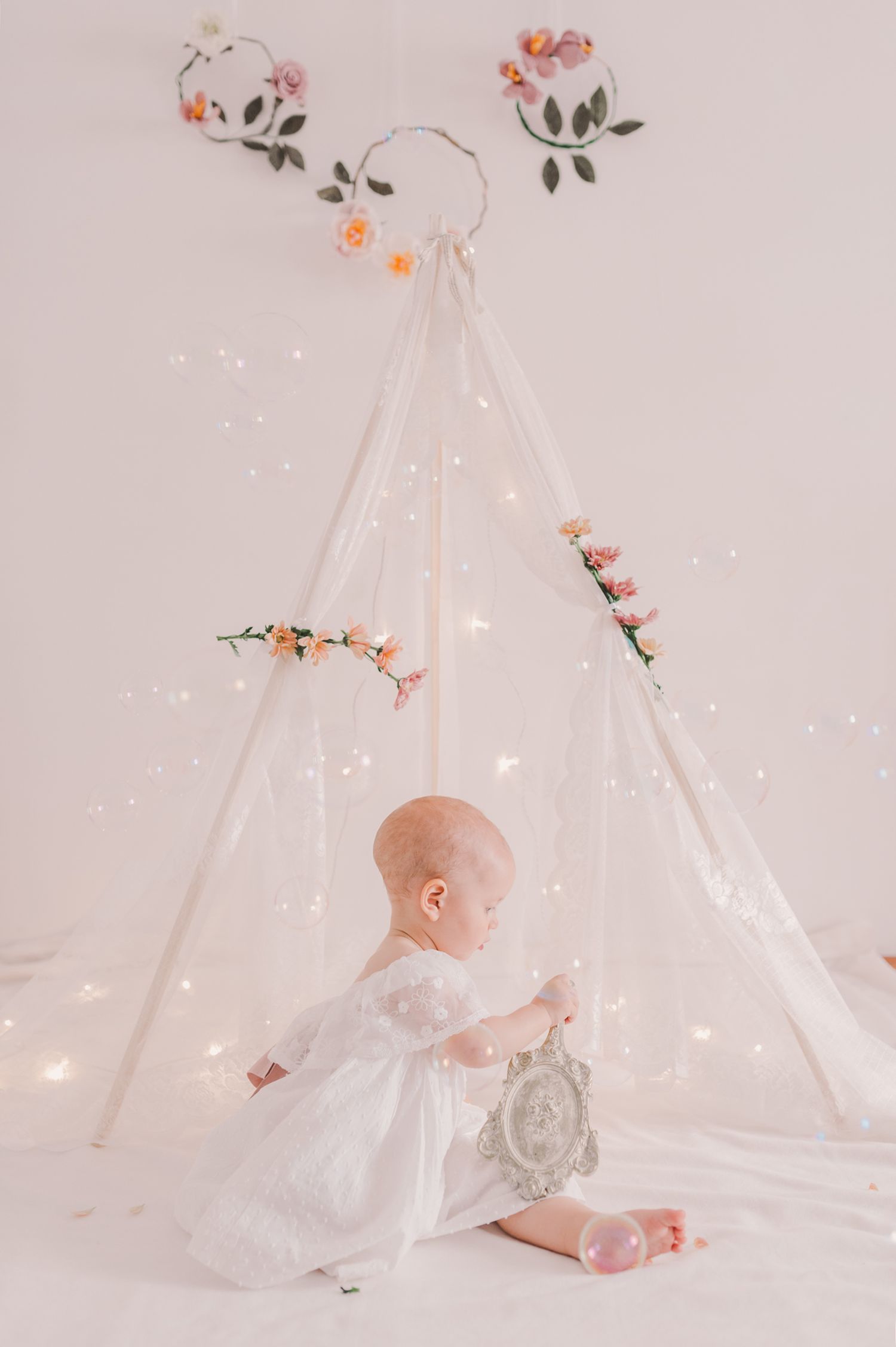 Baby in white dress explores magical teepee setting with twinkling lights and flowers.