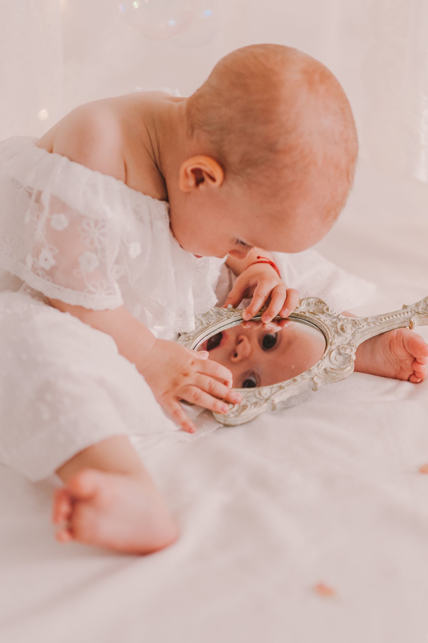 Baby in white dress peering into ornate vintage mirror on light backdrop.