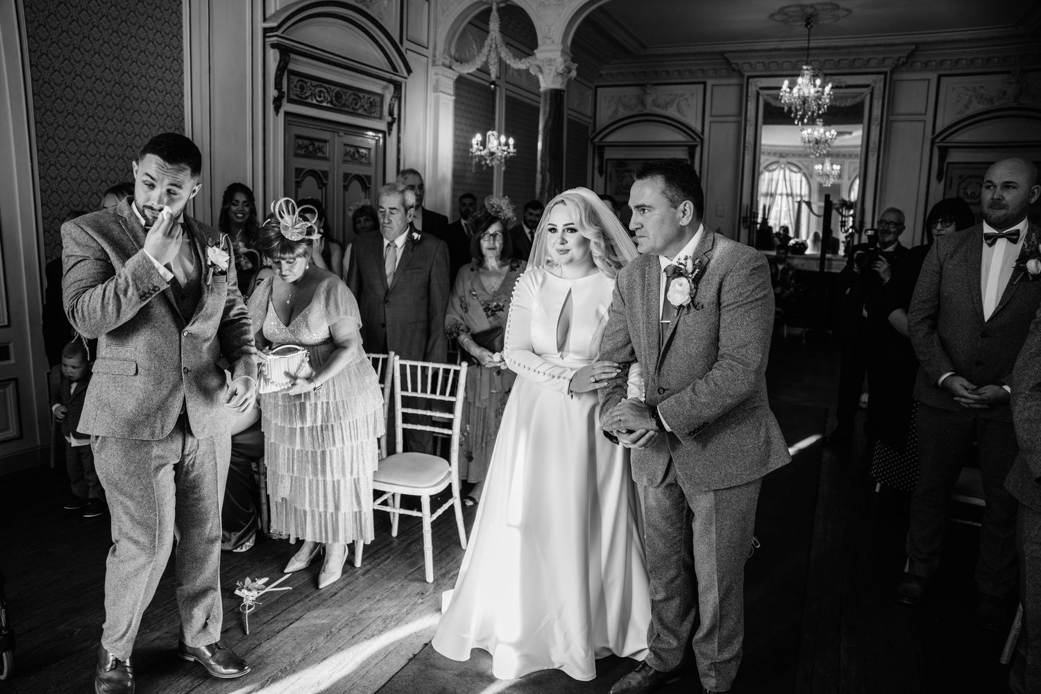 Elegantly dressed wedding guests observe a couple during a ceremony in a grand hall with chandeliers.