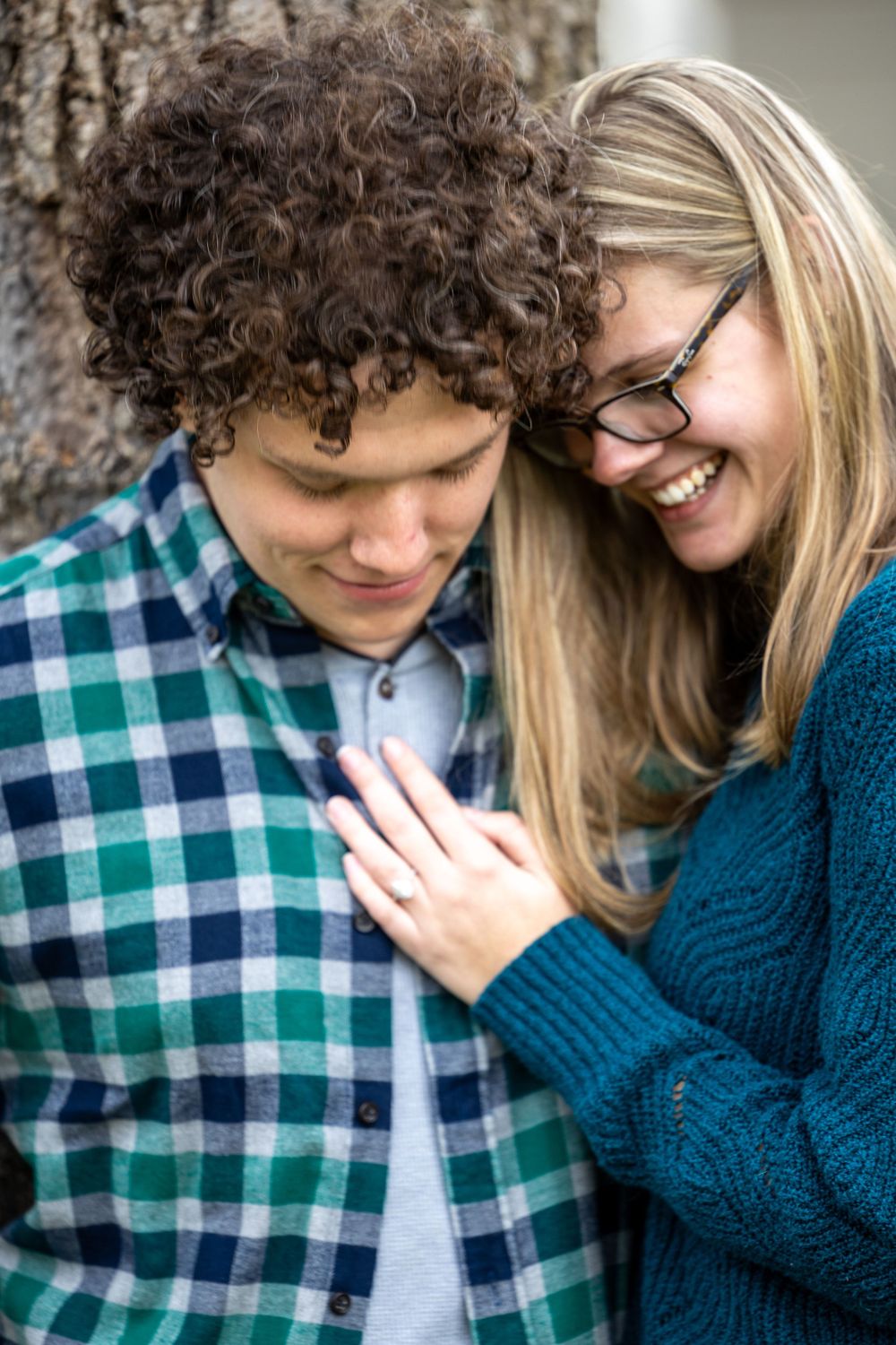 Young couple laughing together near a tree, woman in blue sweater rests hand on man's chest.