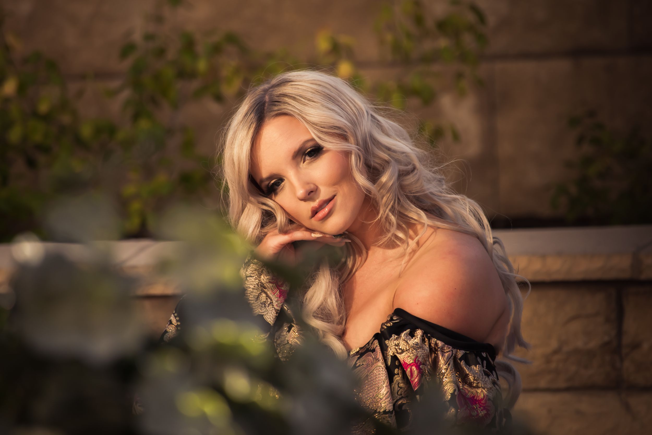 Blonde woman with wavy hair in floral off-shoulder dress poses outdoors at dusk.