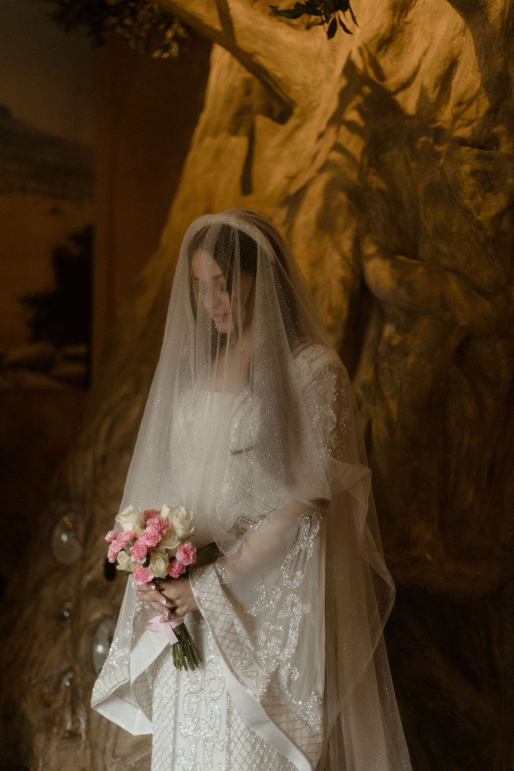 Ethereal bridal portrait with delicate veil and pink bouquet against textured backdrop.