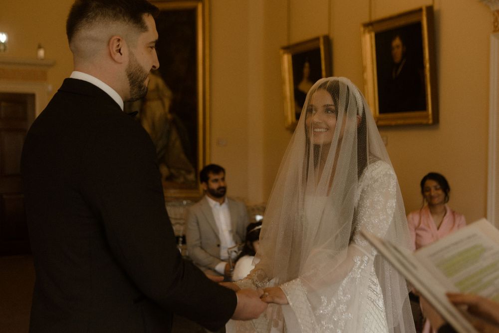 A couple holds hands during their wedding ceremony in a warmly lit historic room.