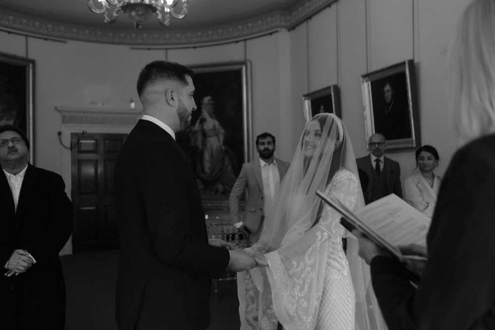 Black and white photograph of a wedding ceremony in an ornate room with historic portraits.