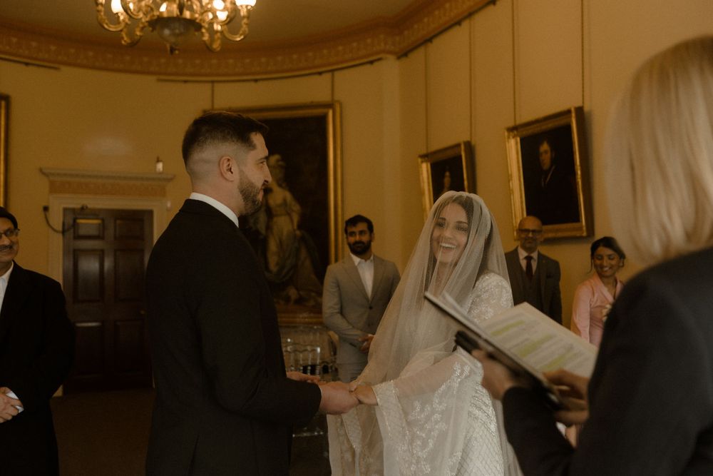 Wedding ceremony in progress in an elegant room with chandelier and historic artwork on walls.
