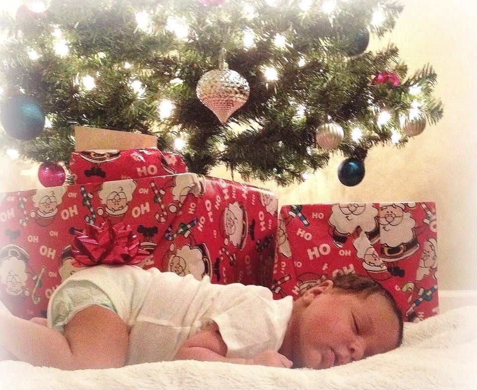 Baby sleeping under Christmas tree surrounded by festive red decorations.