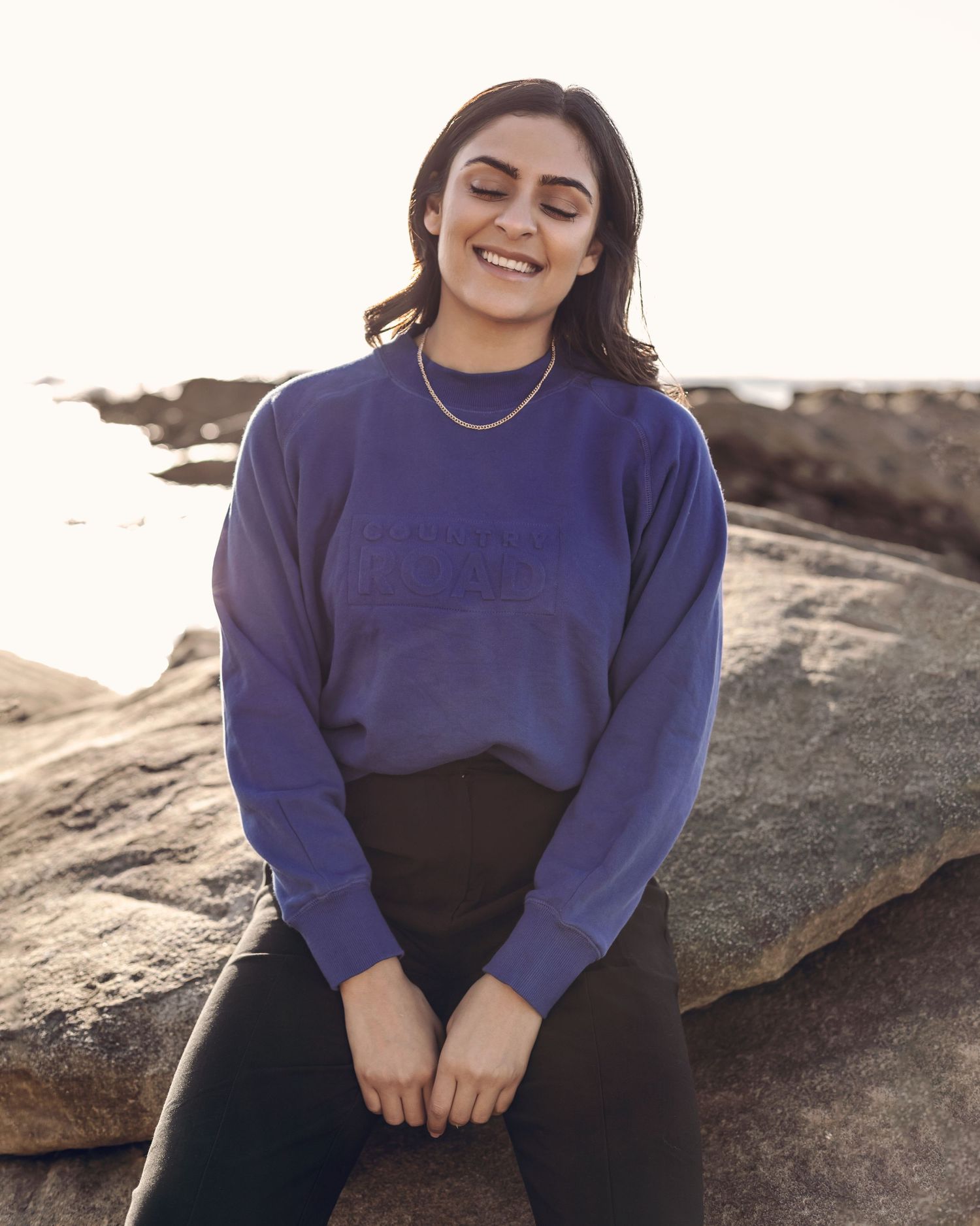 Woman wearing a navy sweater poses on coastal rocks at sunset.