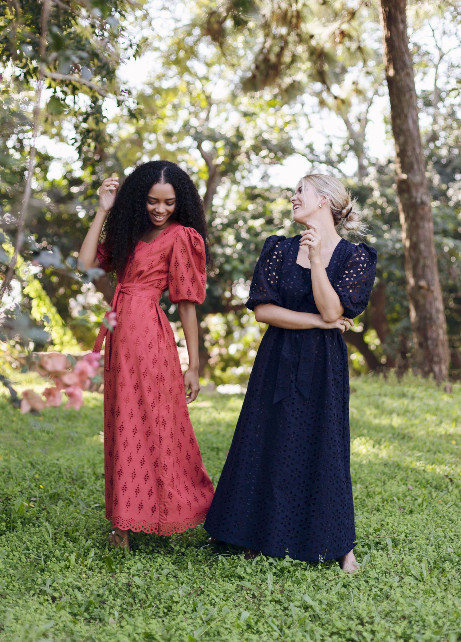 Two women in colorful maxi dresses share a candid moment outdoors surrounded by trees and greenery.