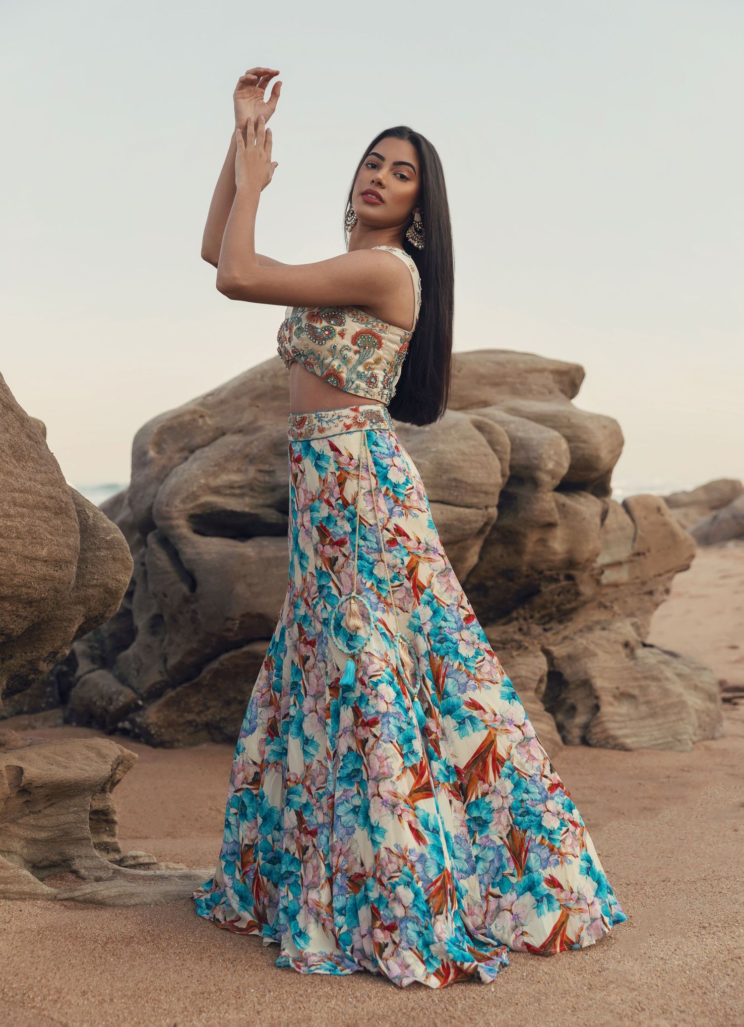 Woman in floral crop top and matching skirt poses dramatically against desert rock formation.