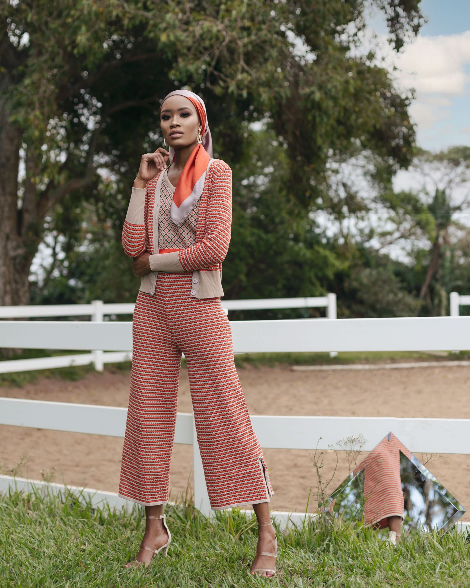 Woman in coral striped pantsuit and headscarf poses elegantly against white fence outdoors.