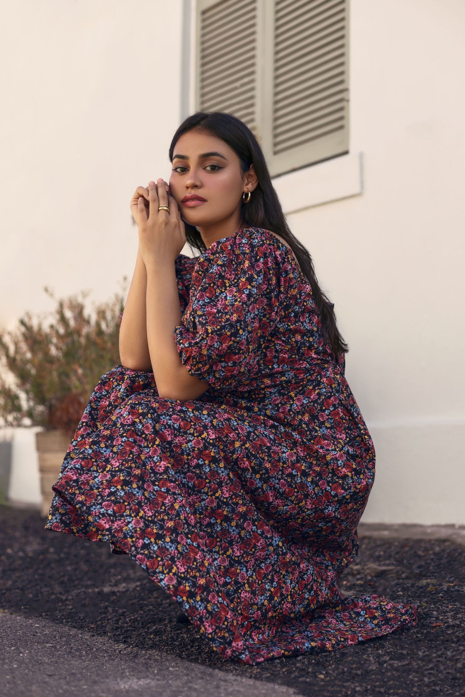 Model wearing floral print maxi dress poses outdoors against white wall.