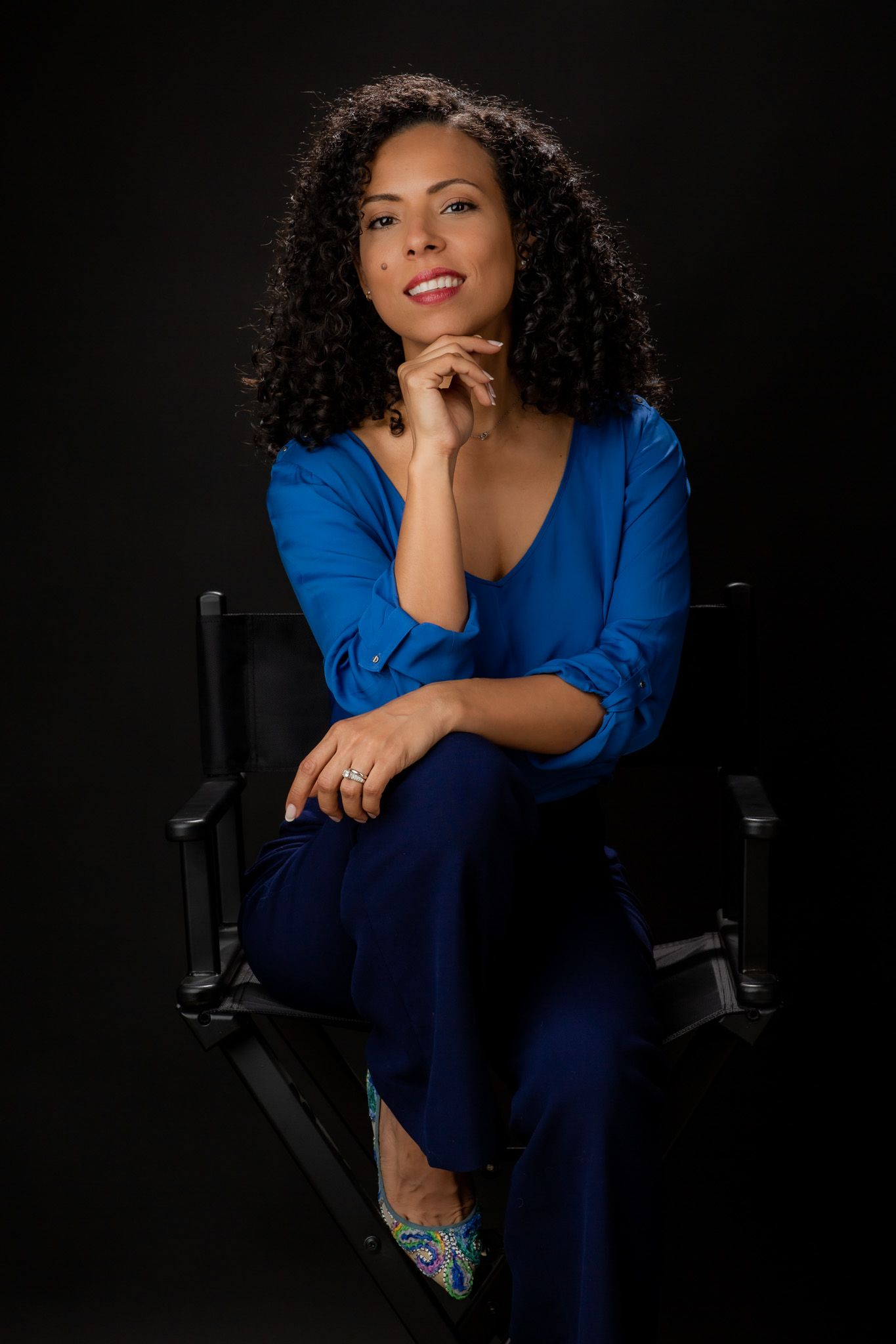 Kelly Montoya during her corporate headshot wearing blue shirt and sitting on a director chair.