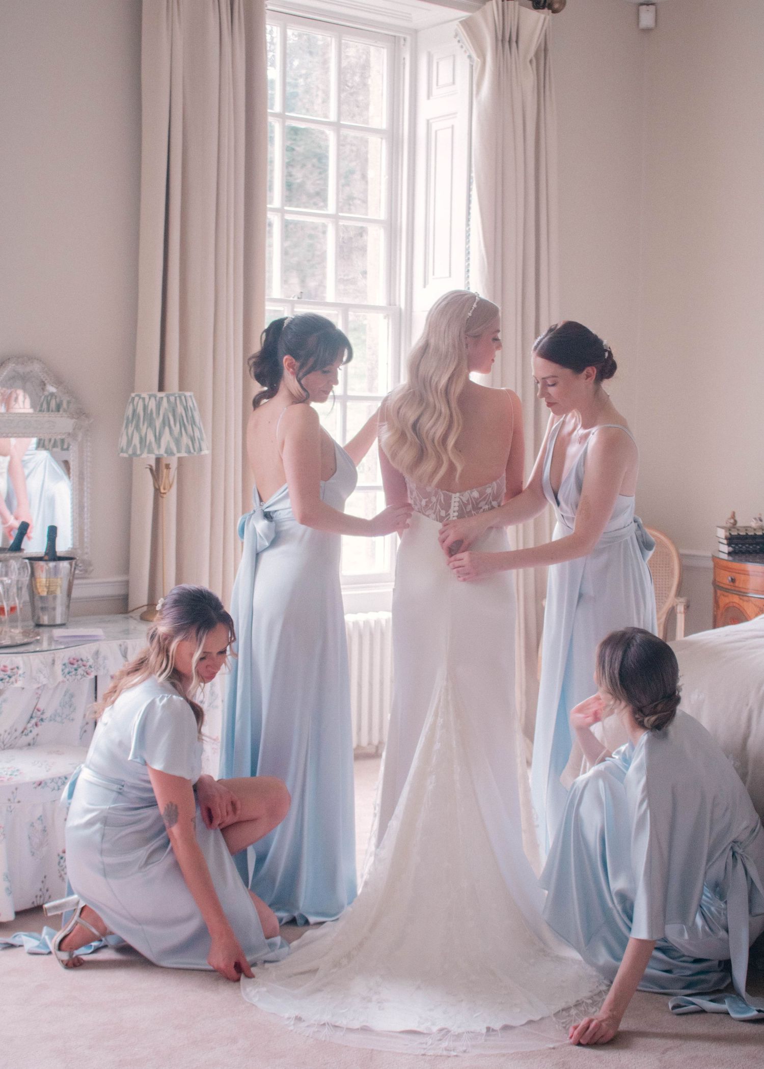 Wedding preparation scene with bridesmaids in light blue dresses helping bride with her gown.