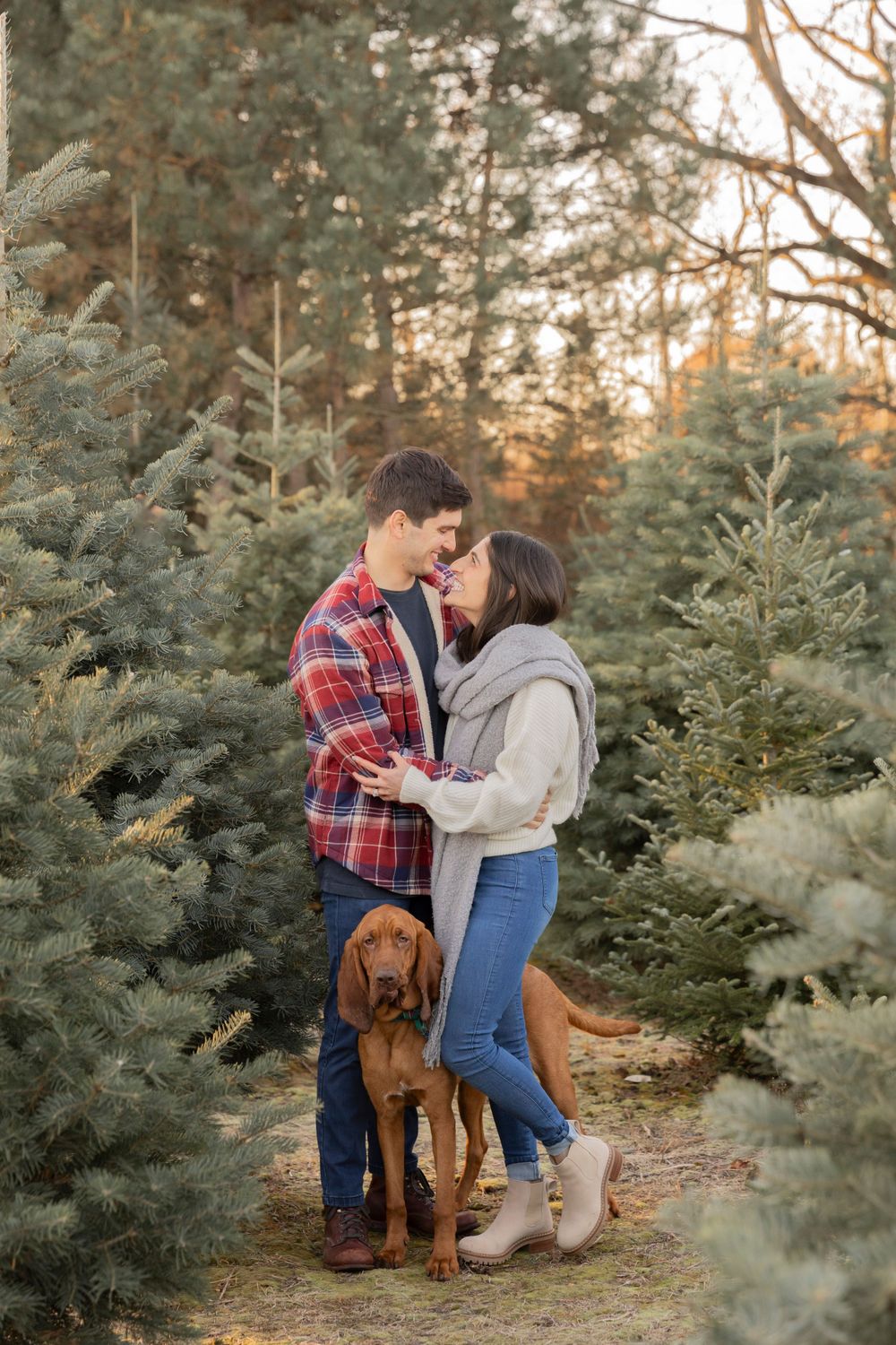 couple in christmas clothes with their puppy smiling at each other at a christmas tree farm