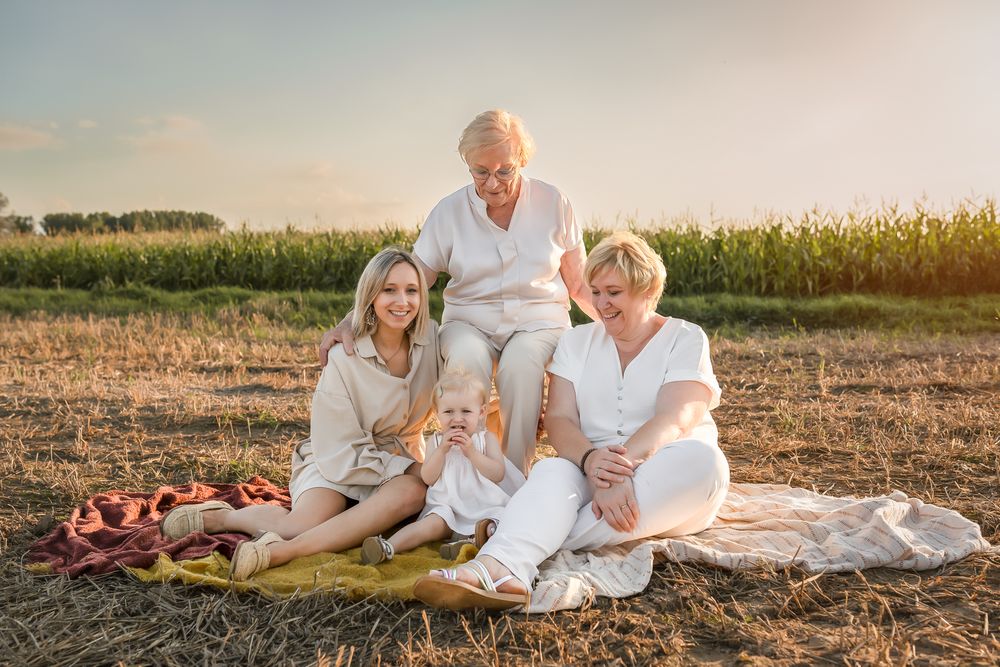A multigenerational group enjoys a sunset picnic on a blanket near a cornfield wearing coordinated white outfits.