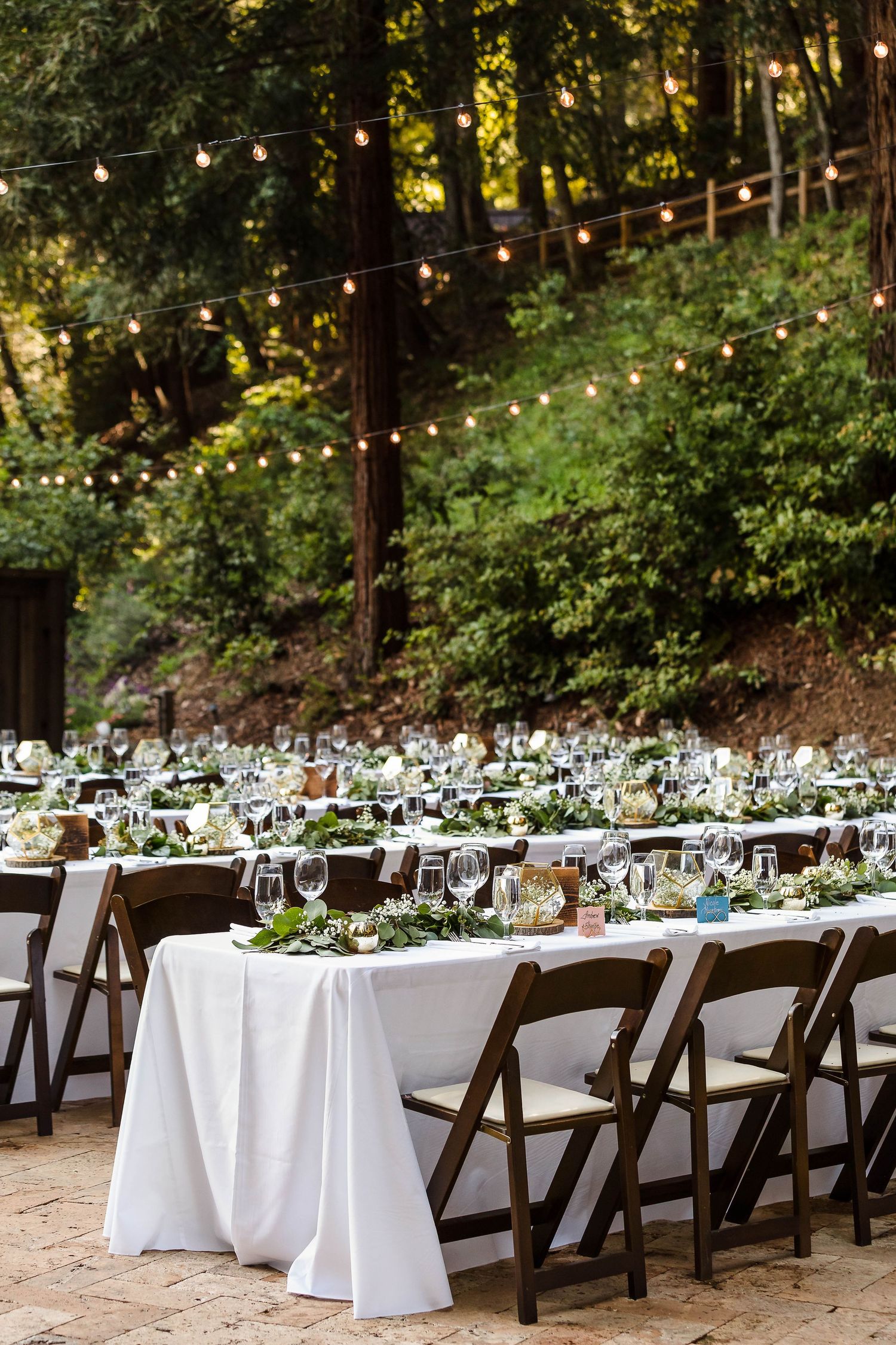 Outdoor forest wedding reception with white tablecloths, wooden chairs, and string lights overhead.