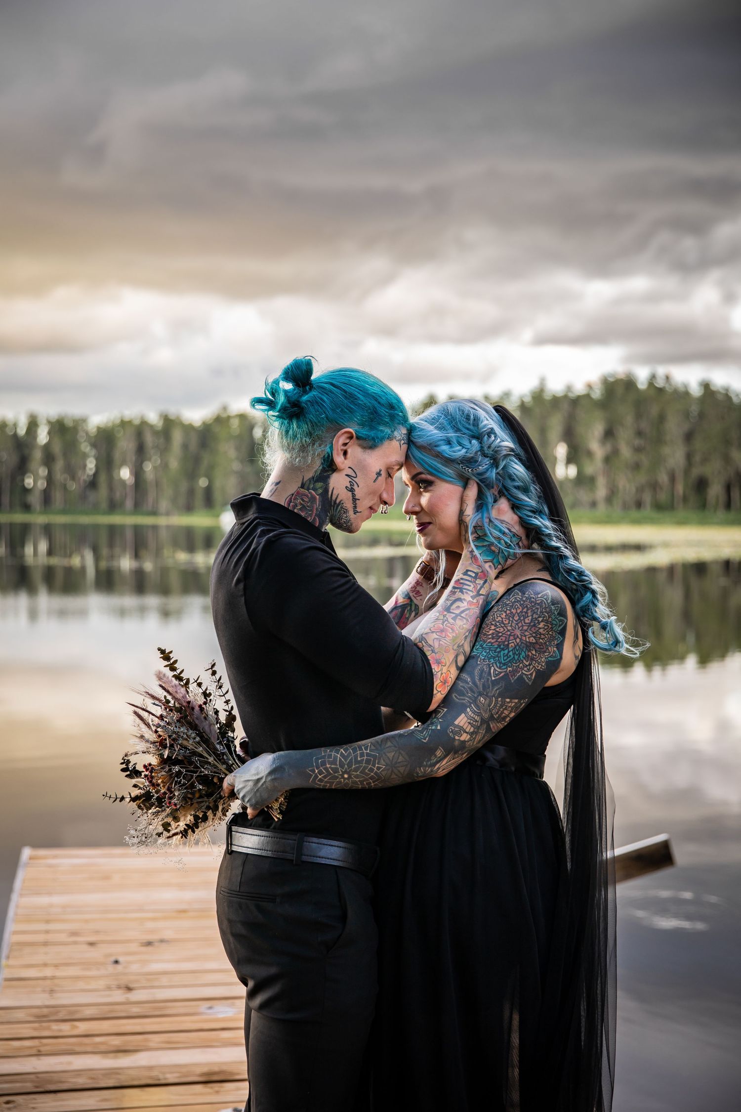 Alternative couple shares intimate moment on dock by lake with colorful hair and dark clothing at sunset.