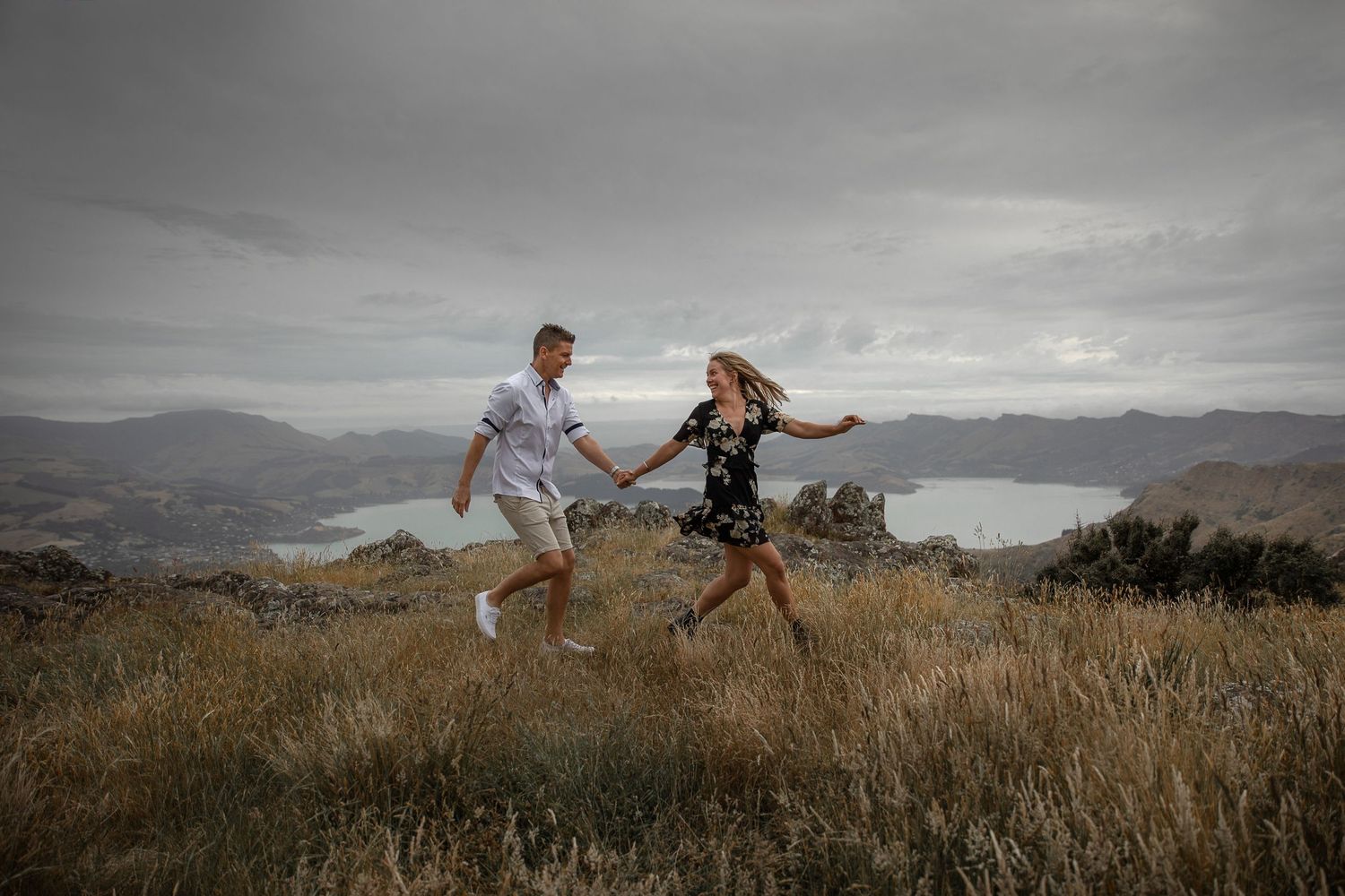 Playful couple jumping together on hilltop with mountain lake view.