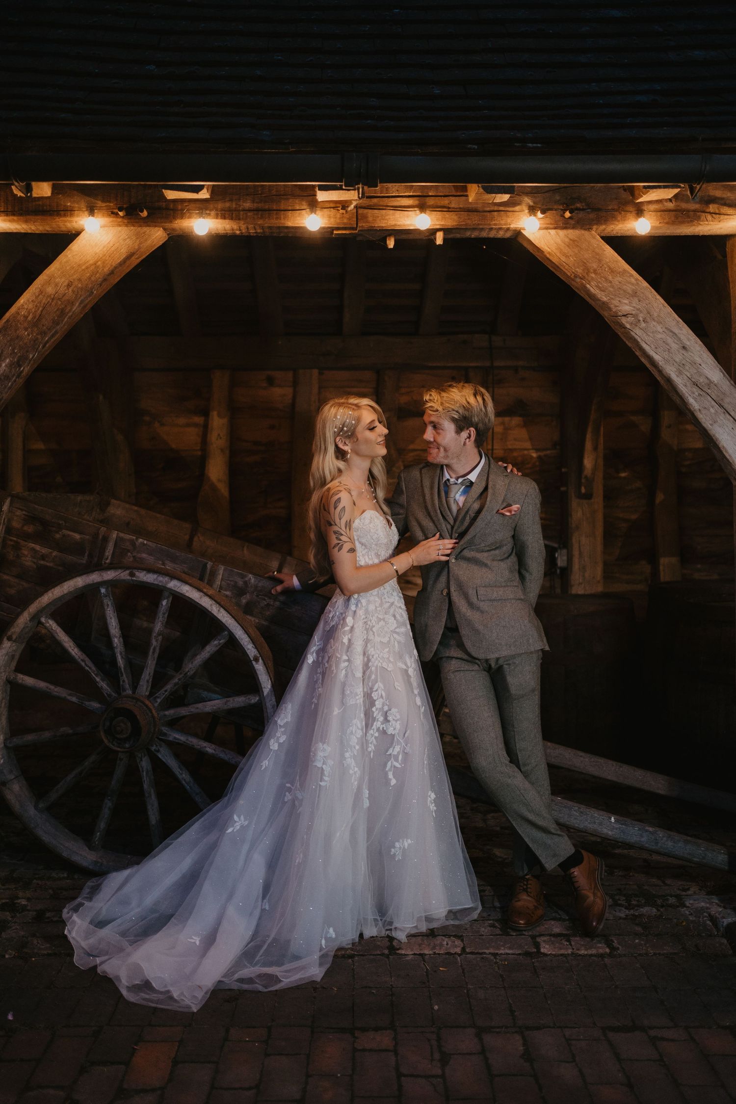 Bride and Groom standing close together at dusk under festoon lights in a barn.