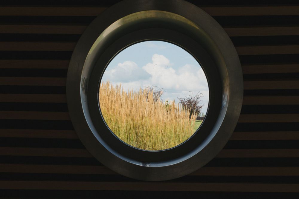 Round porthole window framing golden grass field and blue cloudy sky.