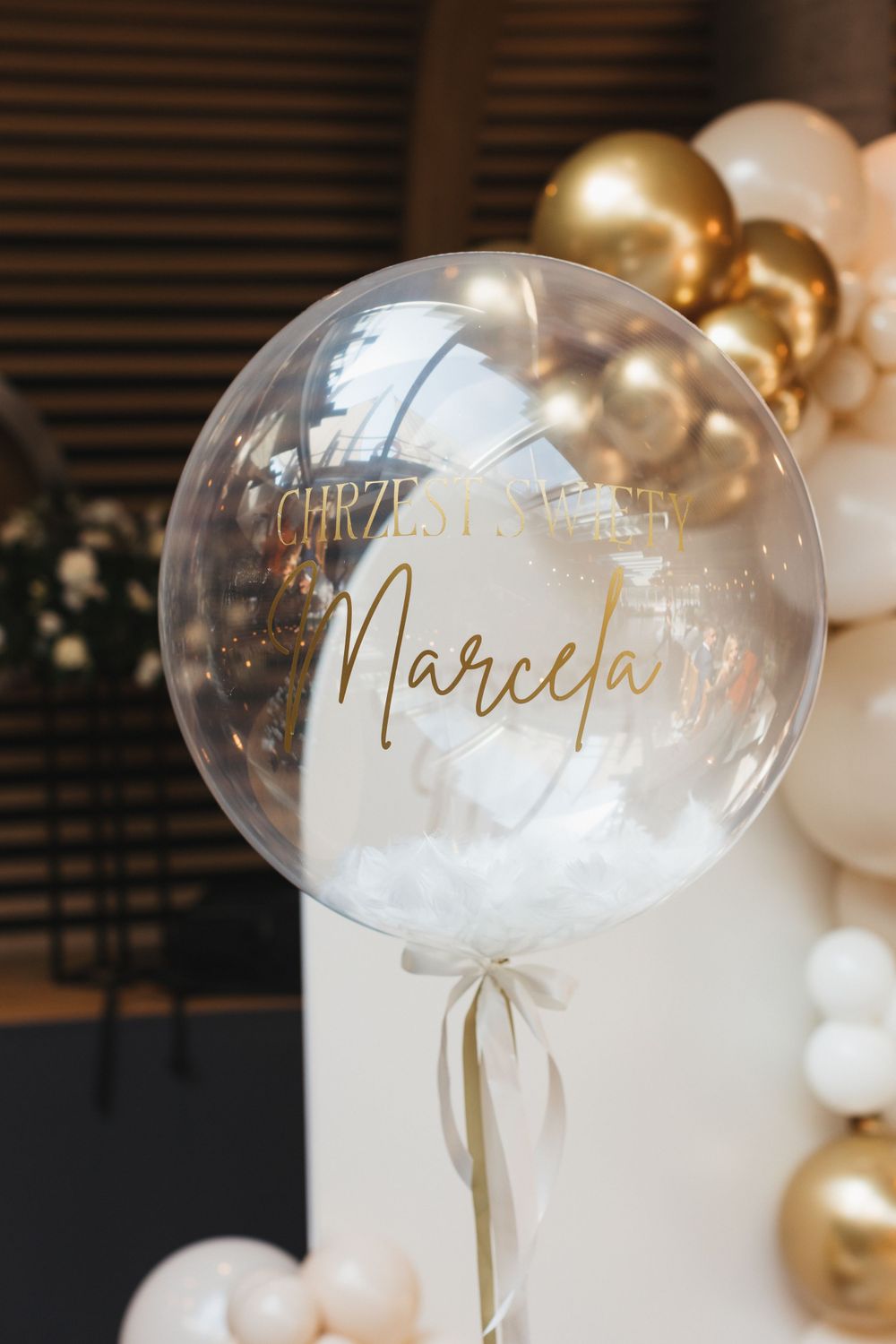 Elegant clear balloon with gold Marcela christening text and white feathers.