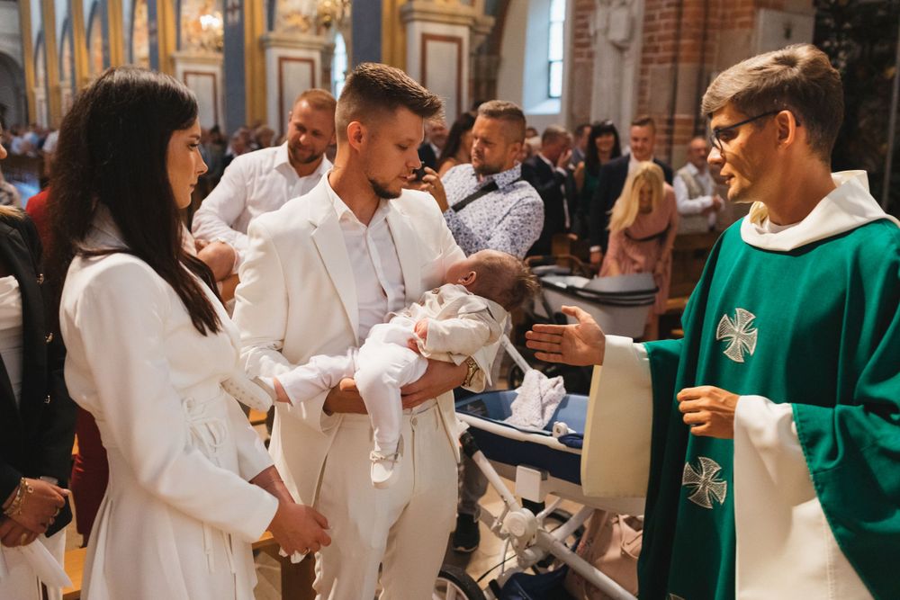 Priest in green vestments performs Catholic baptism ceremony with parents holding infant in white christening gown.