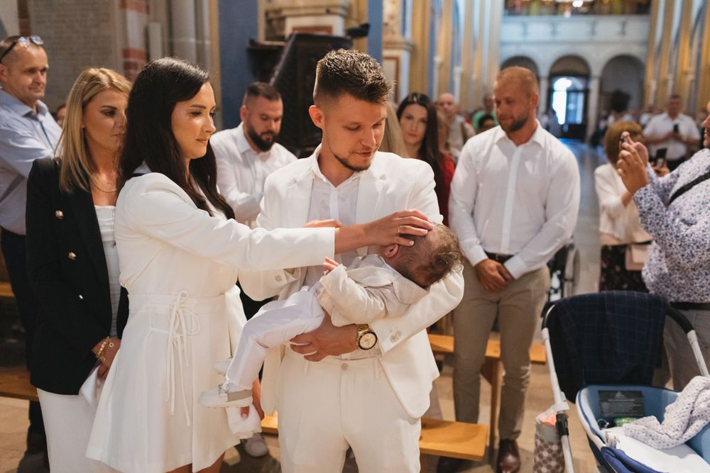 Young couple dressed in white hold their newborn baby during a Catholic baptism ceremony in a church.