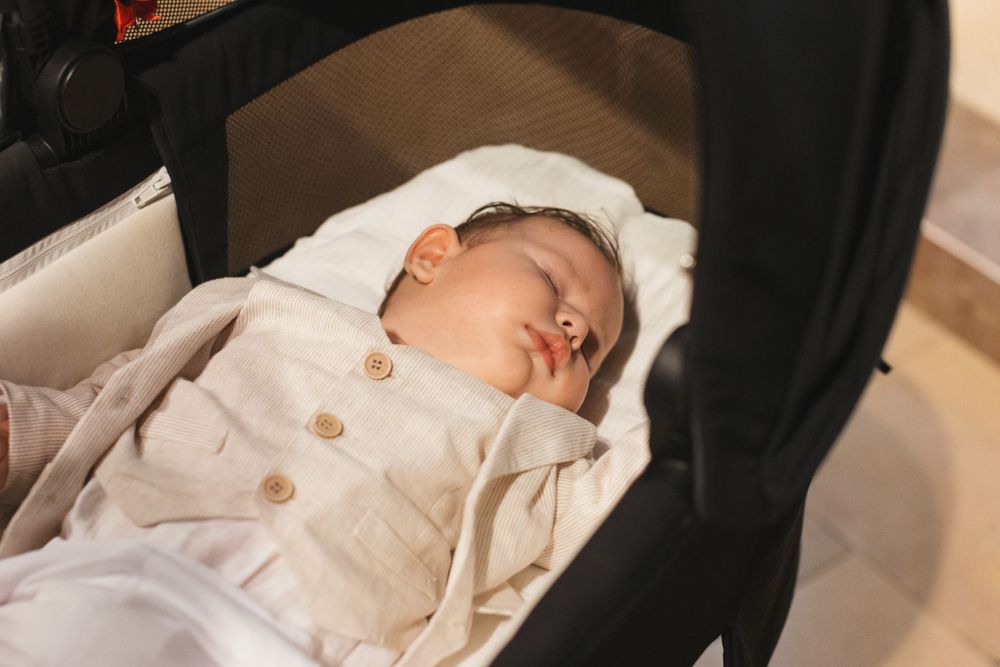 Newborn baby in beige baptism outfit sleeps peacefully in a black pram during christening ceremony.