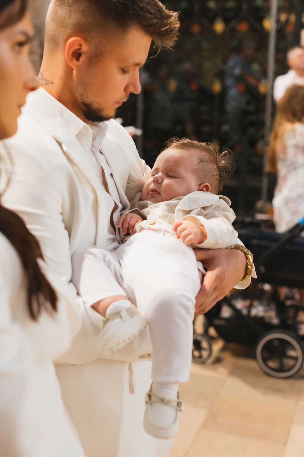 Father in white suit tenderly holds sleeping newborn baby dressed in white during baptism celebration.