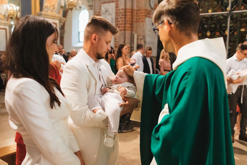 Catholic priest pours holy water over infant during baptism ceremony while parents dressed in white look on.