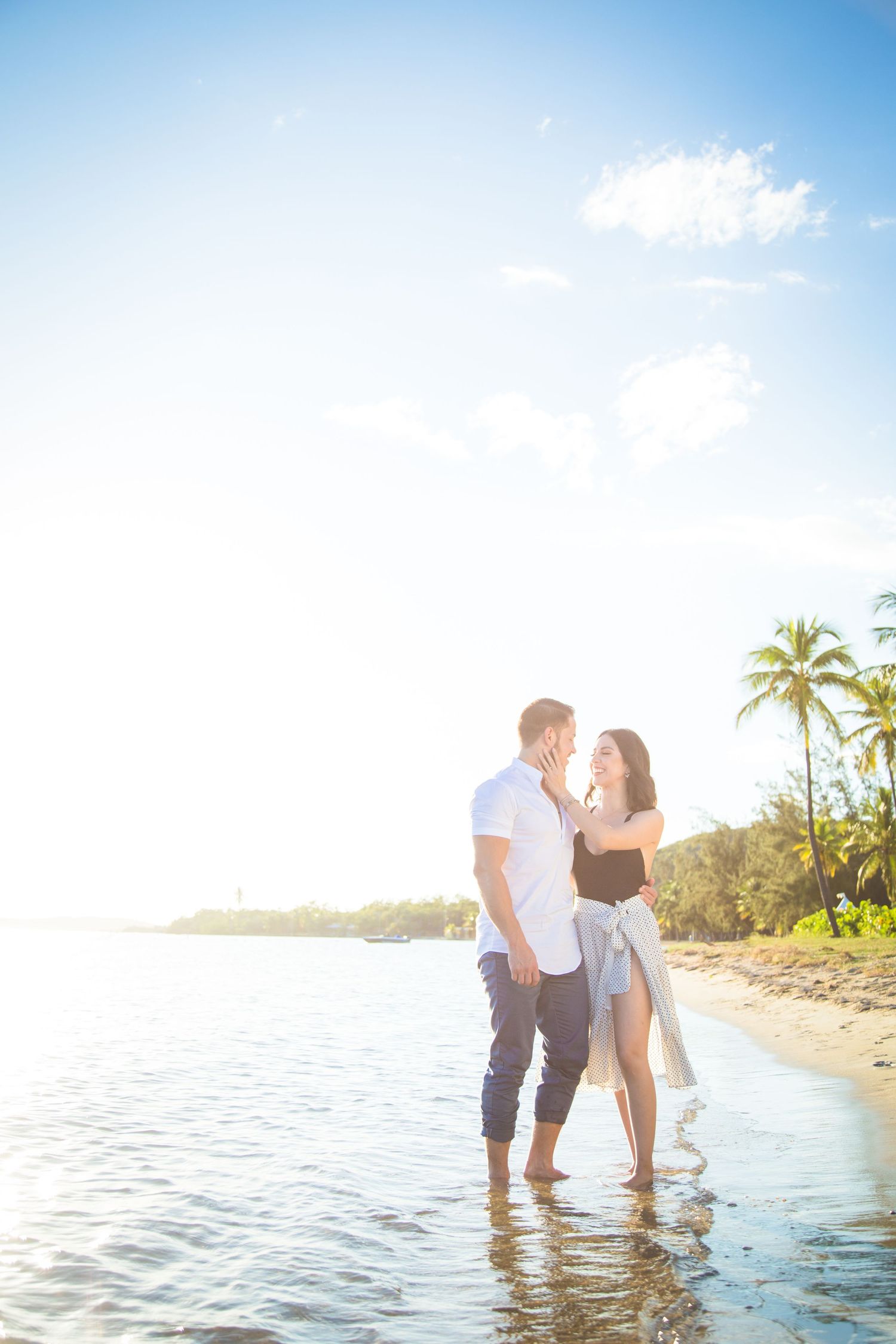 Couple standing in shallow ocean water at sunset with palm trees in background.