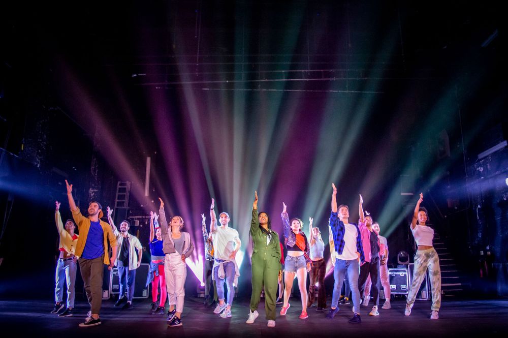 A group of performers raise their arms in unison under colorful stage lights during a theatrical finale.