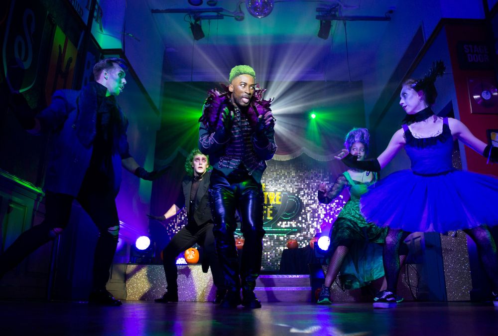 Performers dance in colorful costumes under dramatic stage lighting with green and blue effects.