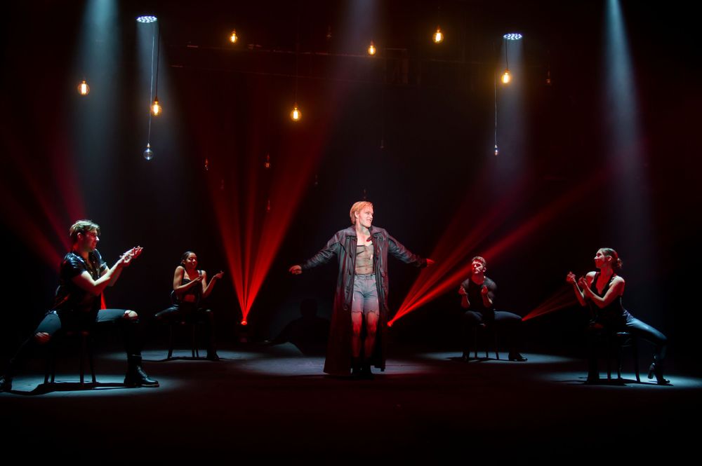 Theater performers on stage with dramatic red lighting and spotlights creating a powerful atmosphere.