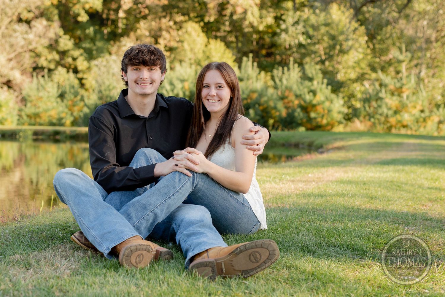 A couple sits together on grass in a romantic outdoor photo session with natural lighting and greenery in background.