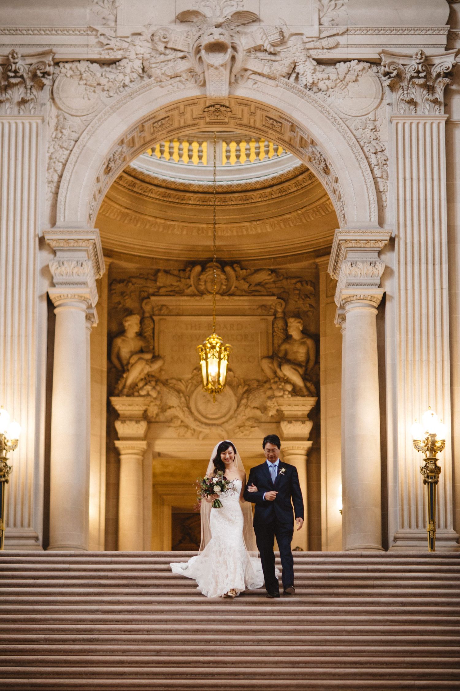 A couple stands at the bottom of an ornate marble staircase beneath a decorated archway.