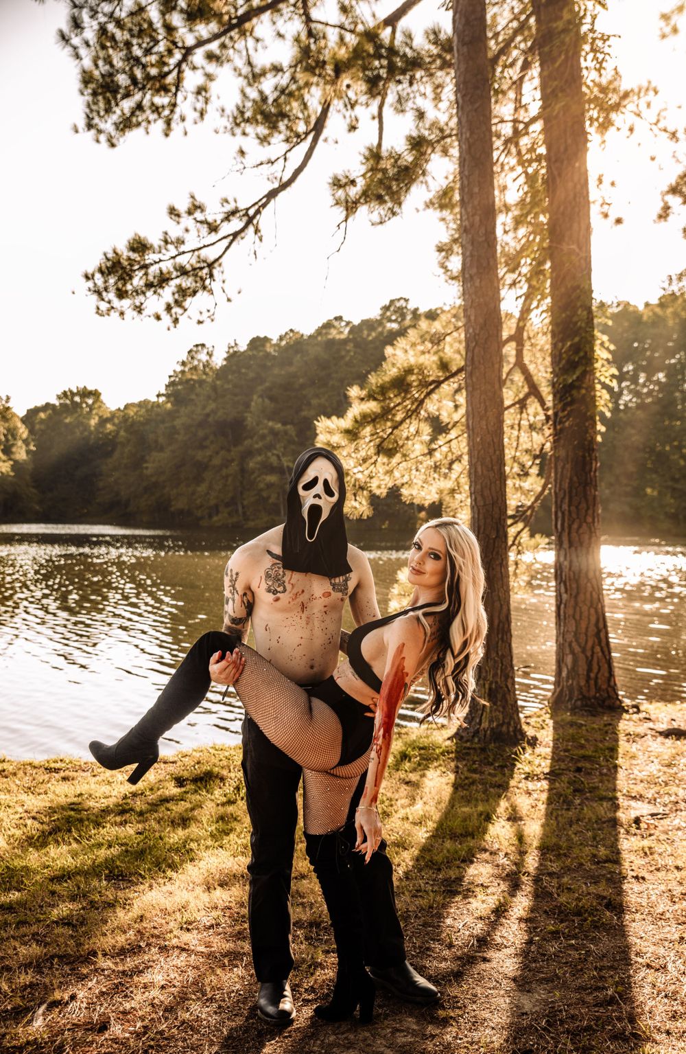 Two figures in Halloween costumes pose playfully near trees at sunset by a lake, creating dramatic silhouettes.