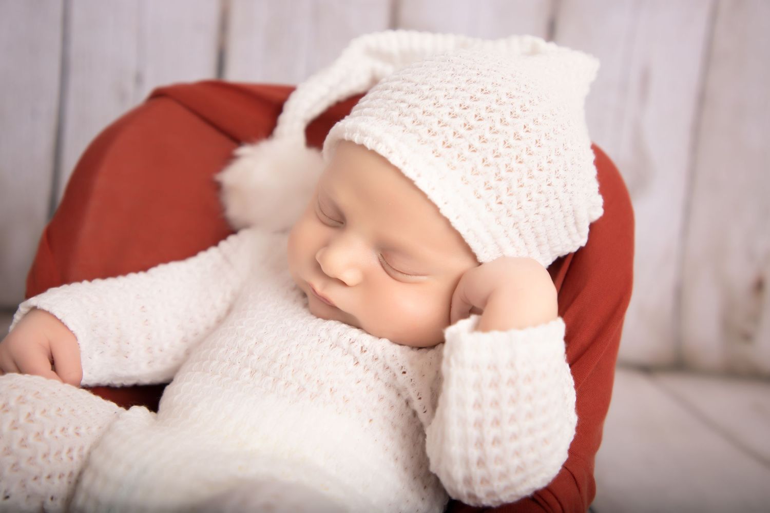 Sleeping baby wearing white knitted outfit and hat in red chair.