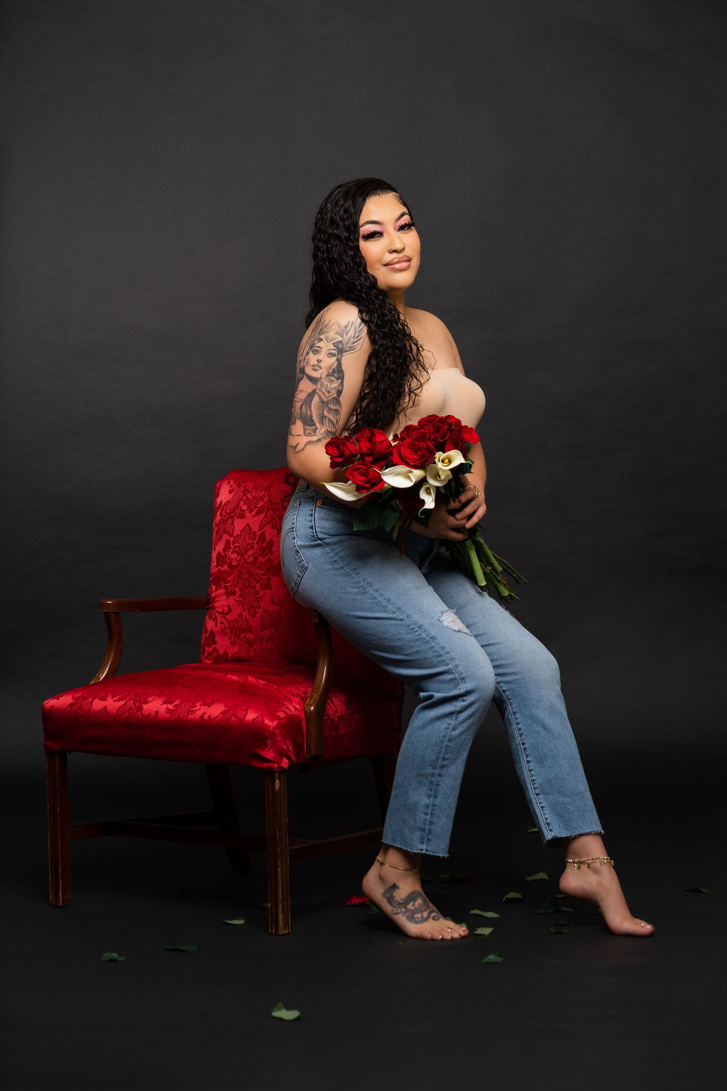 Artistic portrait against black background with person in jeans holding red roses on velvet chair.