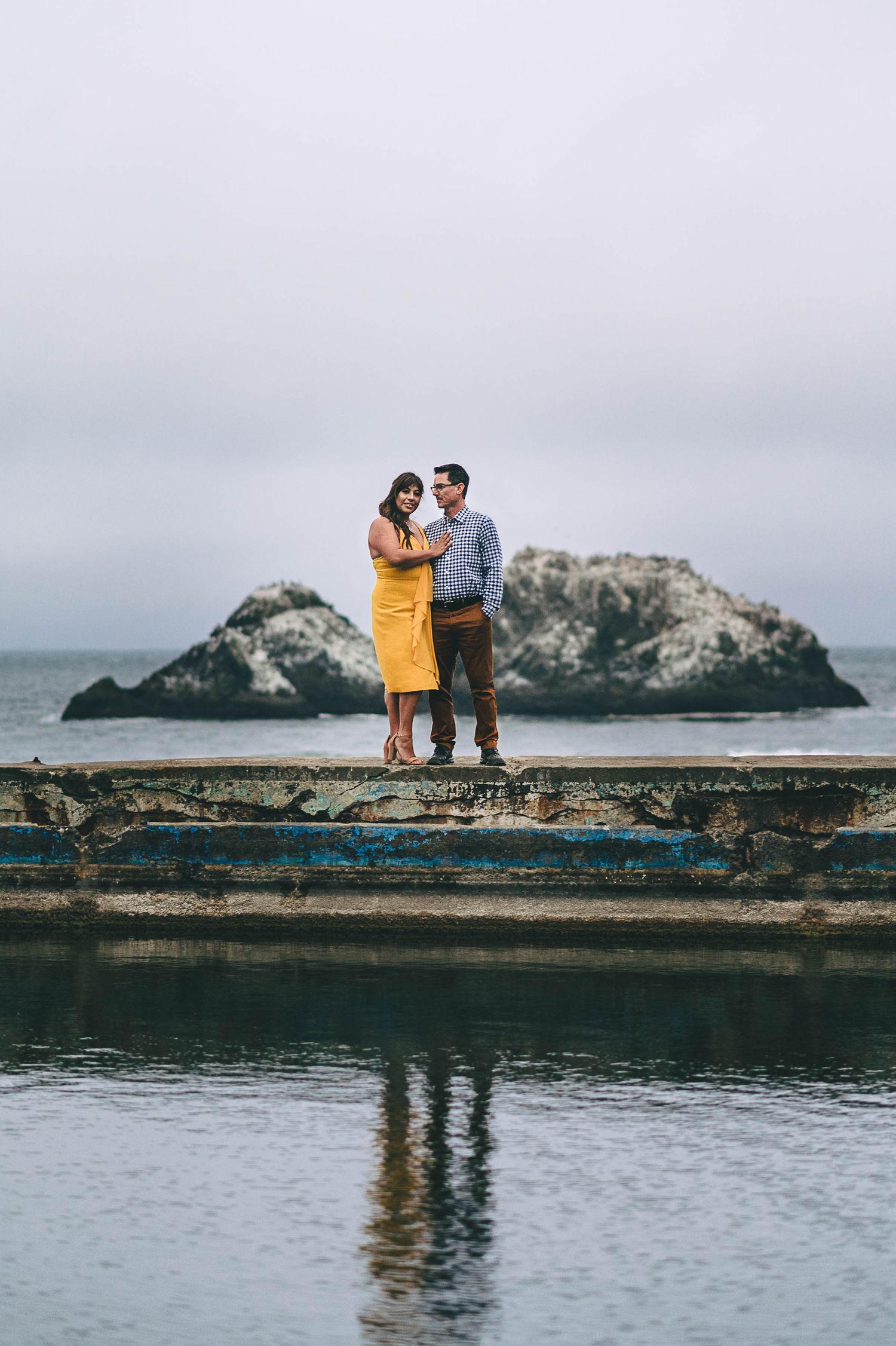 A couple stands on an old pier overlooking a rocky island in the misty ocean.