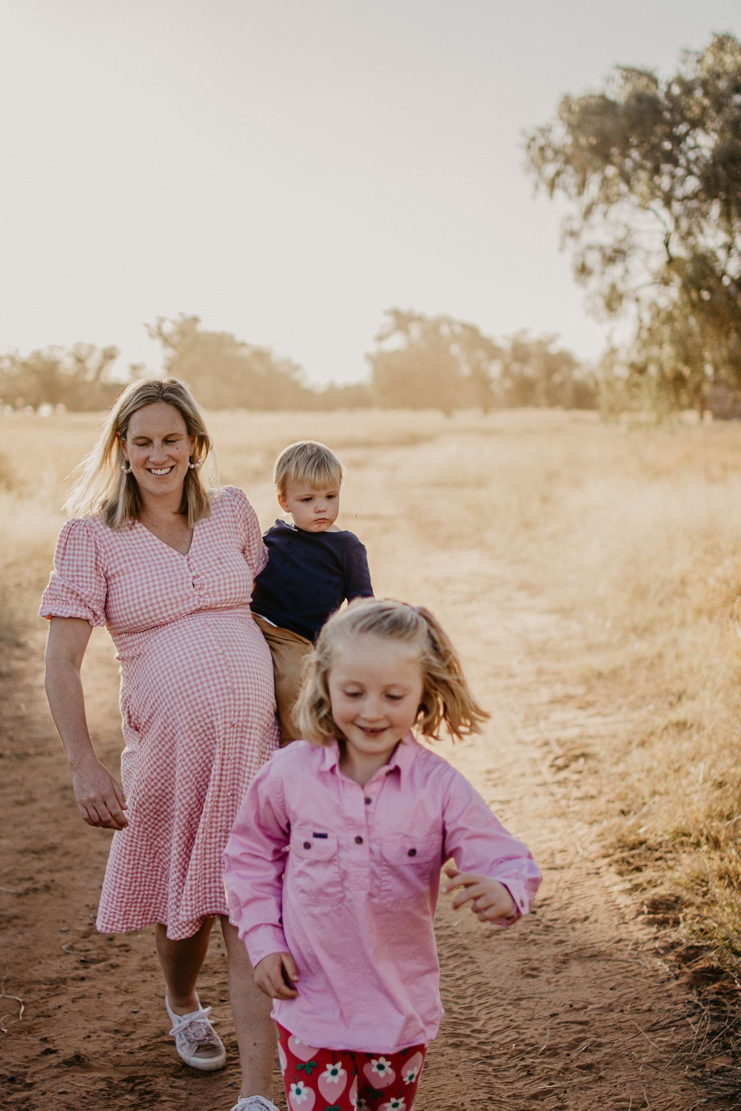 pregnant Mum in a paddock with her kids carrying a toddler on her hip and a another running ahead of them