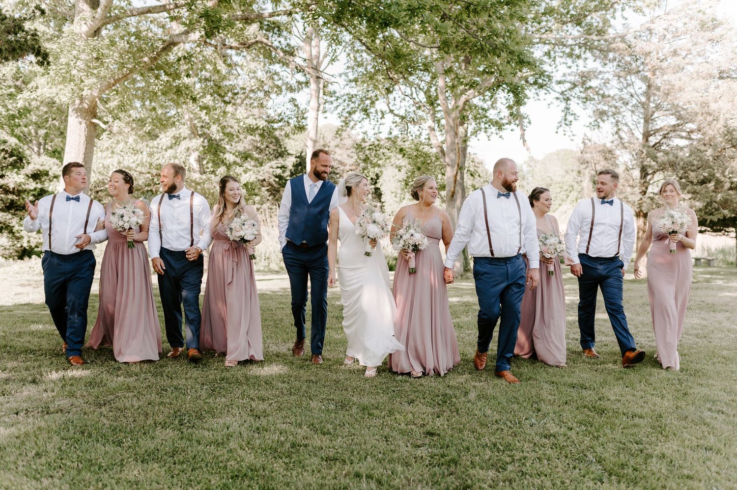 Wedding party in pastel pink dresses and navy suits pose together on a grassy lawn.