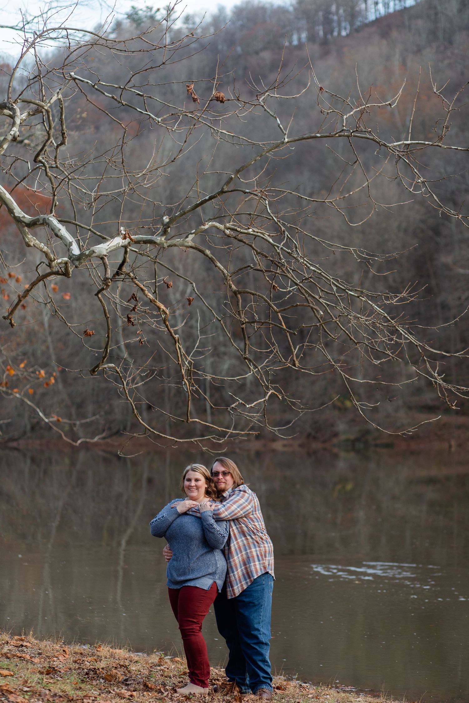 A couple embraces by a serene lake surrounded by bare winter trees and branches.